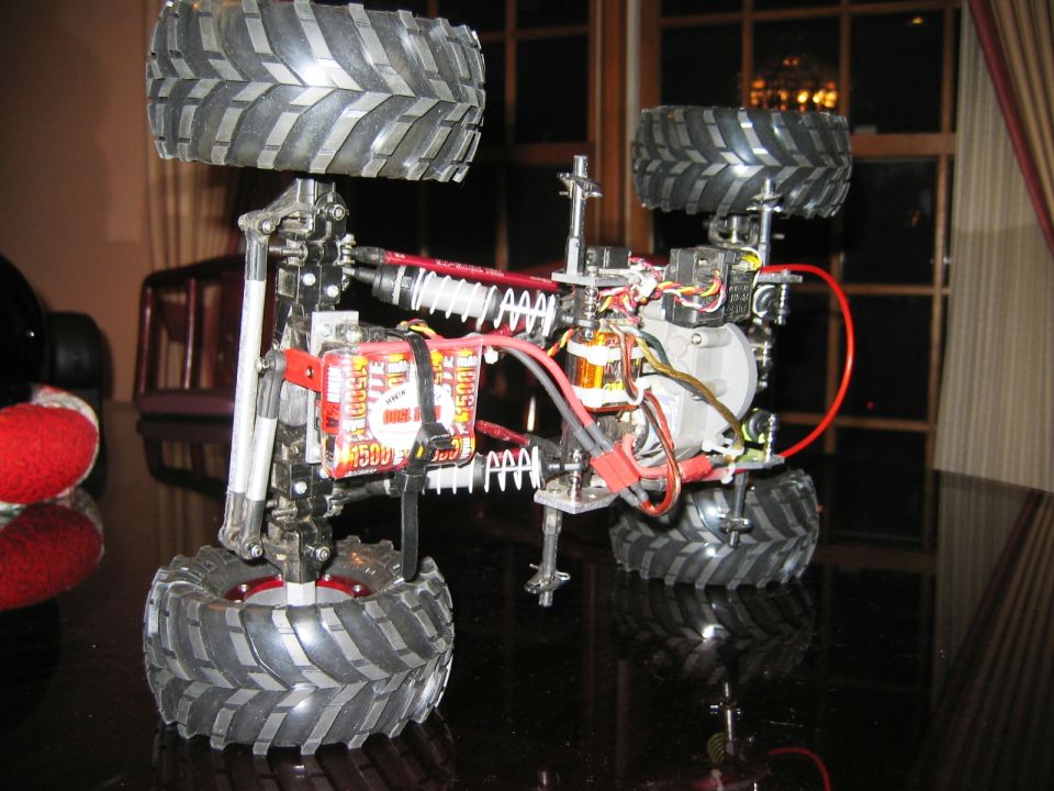 RC off-road car chassis with knobby tires, red frame, electric motor and battery on glass table