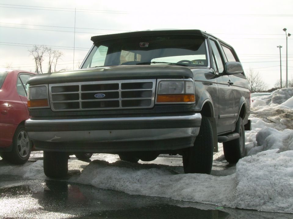 Dark green Ford Bronco climbing a snow pile in a wet parking lot, low front three-quarter view