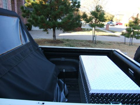 1991 Ford Bronco - The bed of a pickup truck is shown with a soft top and a metal toolbox, with a residential street visible in the background.