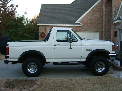 1991 Ford Bronco - A white 1991 Ford Bronco is parked in front of a brick house, showing its side profile and spare tire.