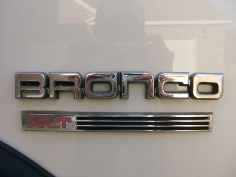 1991 Ford Bronco - Close-up of the chrome 'BRONCO' fender lettering above a ribbed chrome badge with red 'XLT' script, mounted on a white body panel. The poli…