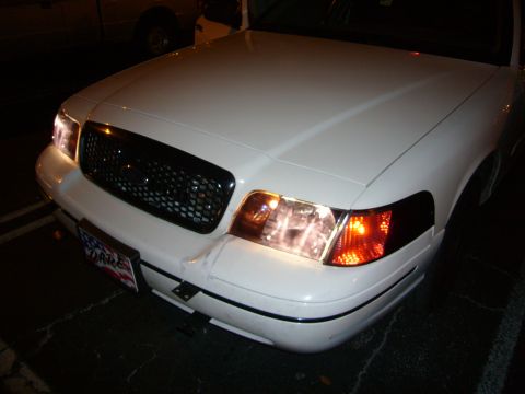 2000 Ford Crown Victoria - The front of a white sedan is shown with its headlights illuminated.