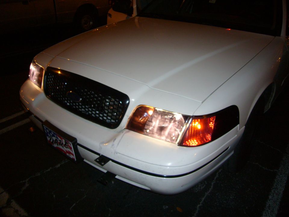 Front of a white sedan with headlights on. License plate says DARE