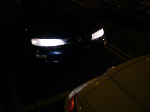 2000 Ford Crown Victoria - The bright headlights of a dark blue sedan are visible at night, with a portion of another vehicle in the foreground.