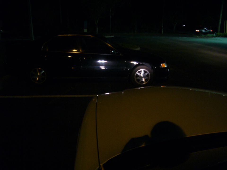 Black sedan parked at night, foreground shows part of another car's hood