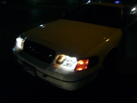 2000 Ford Crown Victoria - The front of a white 2000 Ford Crown Victoria is visible at night, with its headlights illuminated.