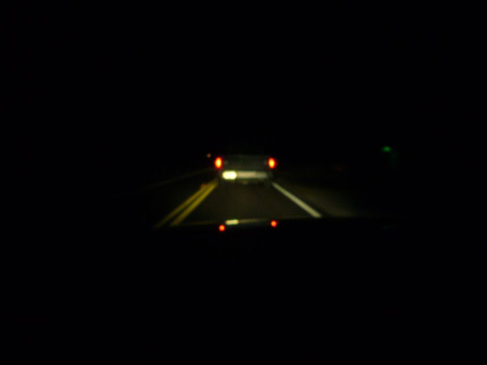Car driving on a dark road at night, seen from inside another vehicle. Taillights of the car ahead are visible