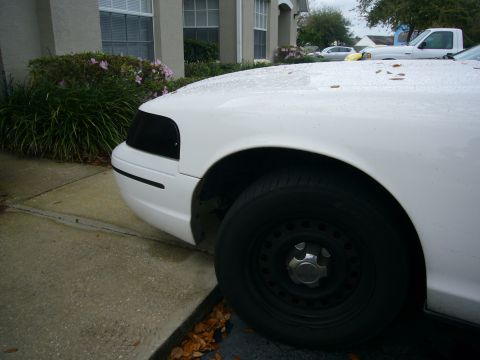 2000 Ford Crown Victoria - The front corner of a white vehicle is shown, a blacked-out headlight and a steel wheel.