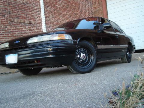 1992 Ford Crown Victoria - Black 1992 Ford Crown Victoria sedan with steel wheels parked in front of a brick building and garage door.