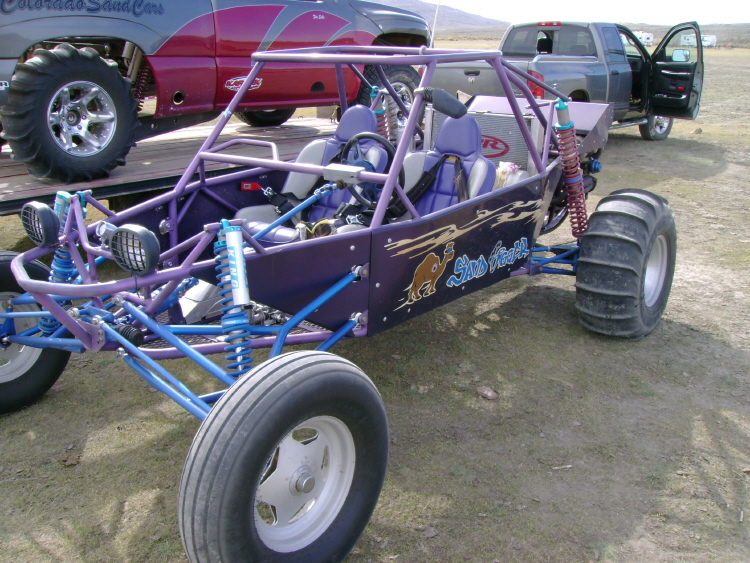 Purple custom sand rail buggy with two seats, sand tires, and a camel graphic on the side. A pickup truck is parked behind it