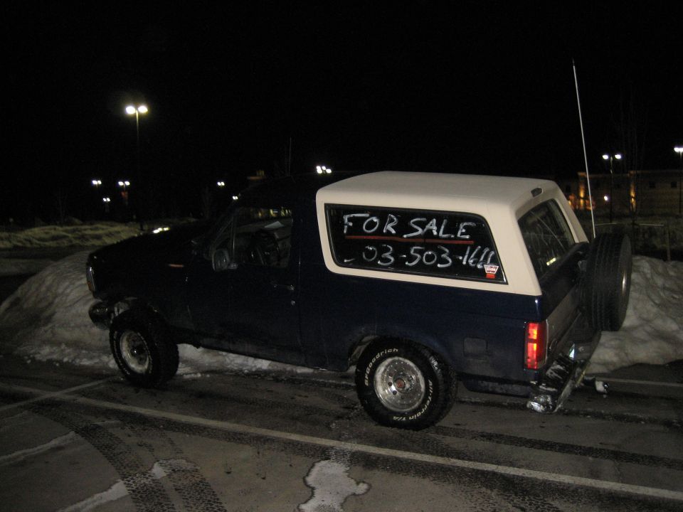 Dark blue Ford Bronco with white top at night, 'FOR SALE' and phone number written on rear window, snow in background