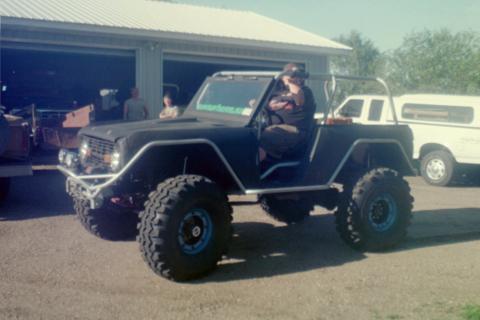 1995 Ford Bronco - This heavily modified SUV features oversized tires and a roll cage, with a pickup truck visible behind it.