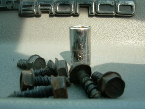 1988 Ford Bronco - A socket and several screws are laid out on a textured surface, with a 'BRONCO' emblem visible in the background.