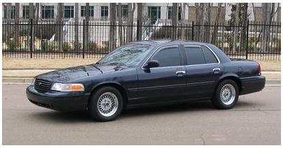 1999 Ford Crown Victoria - A dark blue sedan is parked on the street in front of a building with a black fence.