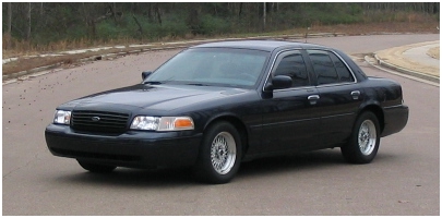 1999 Ford Crown Victoria - A black Ford Crown Victoria sedan is shown from the front-right quarter angle on a paved road with trees in the background.