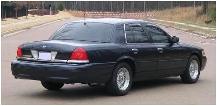 1999 Ford Crown Victoria - Rear view of a black sedan on a paved road with trees in the background.