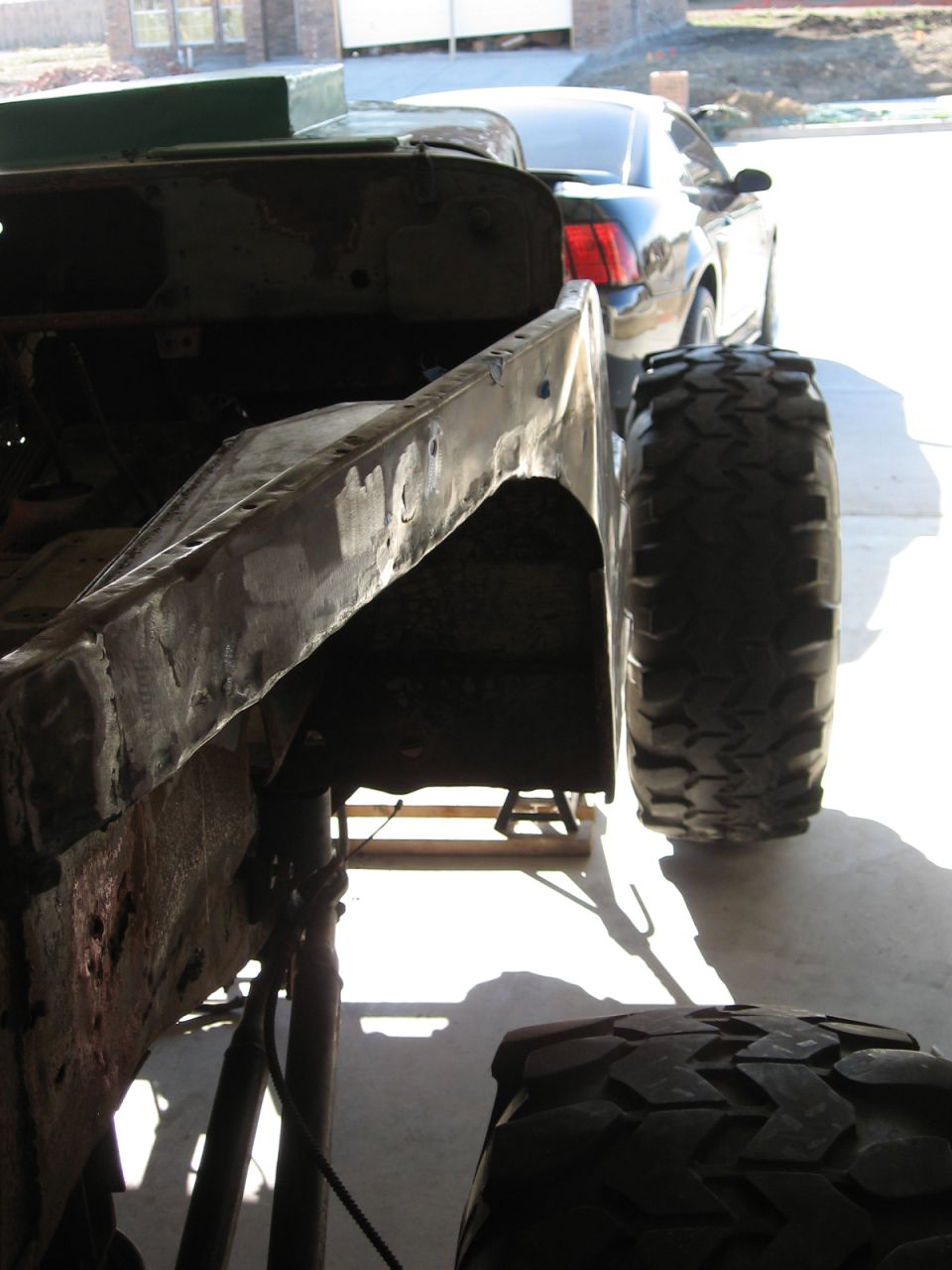 Partially restored vehicle frame and large off-road tire. A modern car is visible in the background
