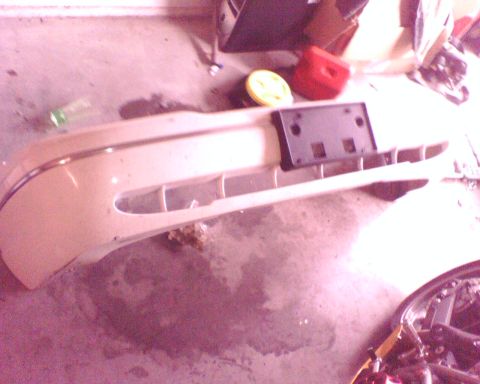 2000 Ford Crown Victoria - Front bumper cover in cream/white paint lying face-up on a garage floor, with its chrome strip along the top edge and a black plastic licen…