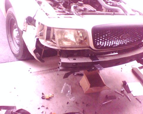 2000 Ford Crown Victoria - Front end of a white Ford sedan mid-disassembly in a garage, with the front bumper cover removed to expose the steel impact bar, brackets a…