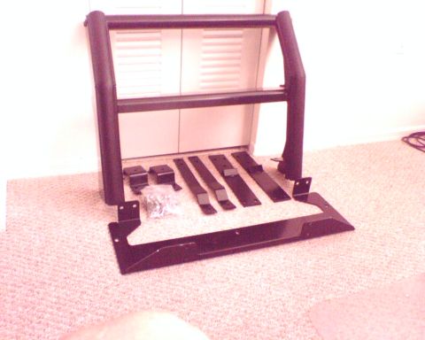 2000 Ford Crown Victoria - A black tubular push bar / grille guard leaning against a wall indoors on carpet, laid out with its full mounting kit. Flat steel mounting…