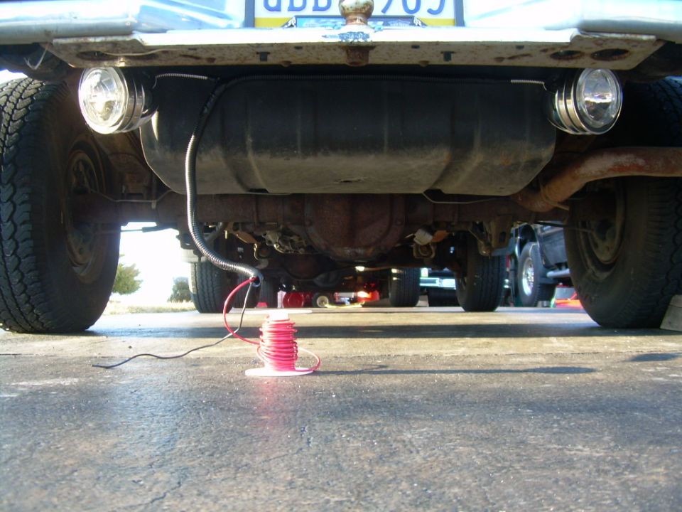 Undercarriage view of a vehicle, showing a fuel tank, exhaust, and wiring. Another vehicle is visible in the background