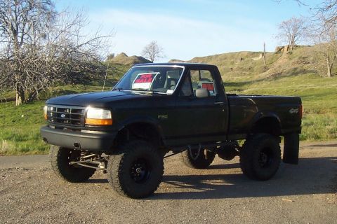 1994 Ford F-150 - A lifted 1994 Ford F-150 pickup truck is parked on a gravel surface with a grassy hill in the background.