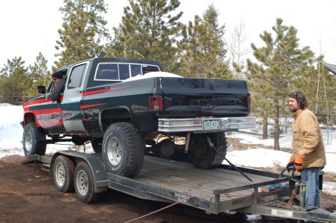 1985 Chevrolet C/K Pickup - A man secures a lifted pickup truck onto a trailer, with snow visible on the ground and trees in the background.
