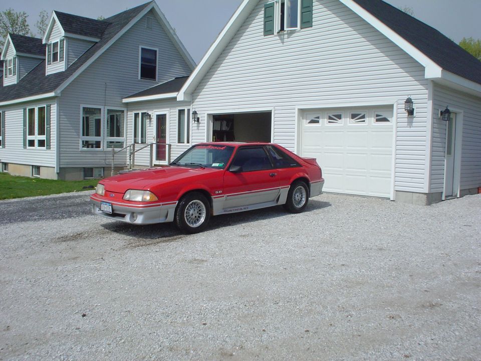 1989 Ford Mustang GT parked on gravel in front of a white garage and house
