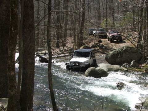 1996 Ford Bronco - A convoy of off-road vehicles navigates a shallow, rocky riverbed in a wooded area.