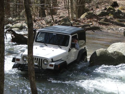 1996 Ford Bronco - White Jeep Wrangler navigates a rocky stream in a wooded area.
