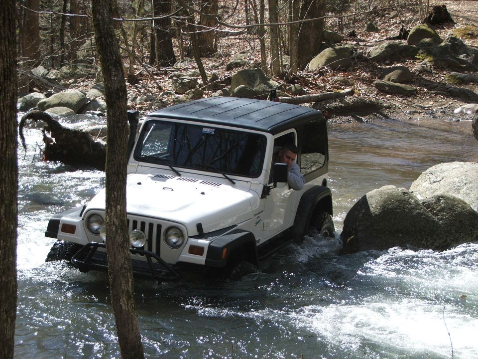 White Jeep Wrangler with black top driving through a rocky stream, driver visible