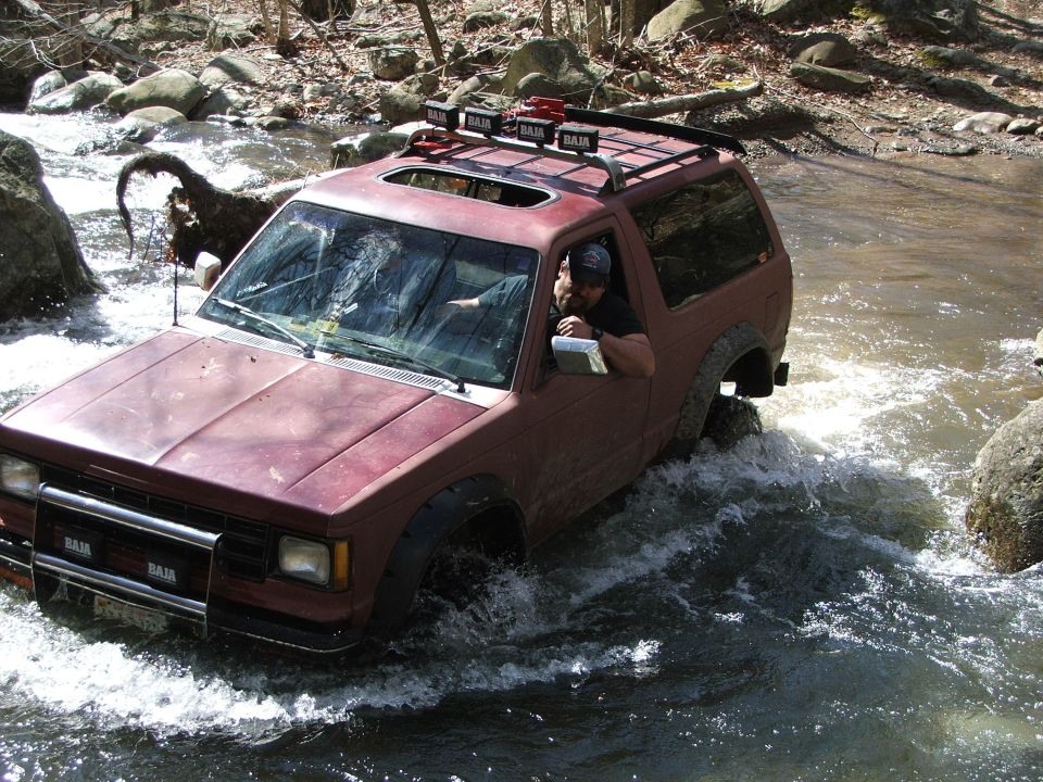 Red SUV with a roof rack driving through a rocky river, driver visible
