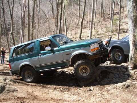 1996 Ford Bronco - A two-tone green and silver 1996 Ford Bronco navigates a rocky trail, with another vehicle visible to its right.