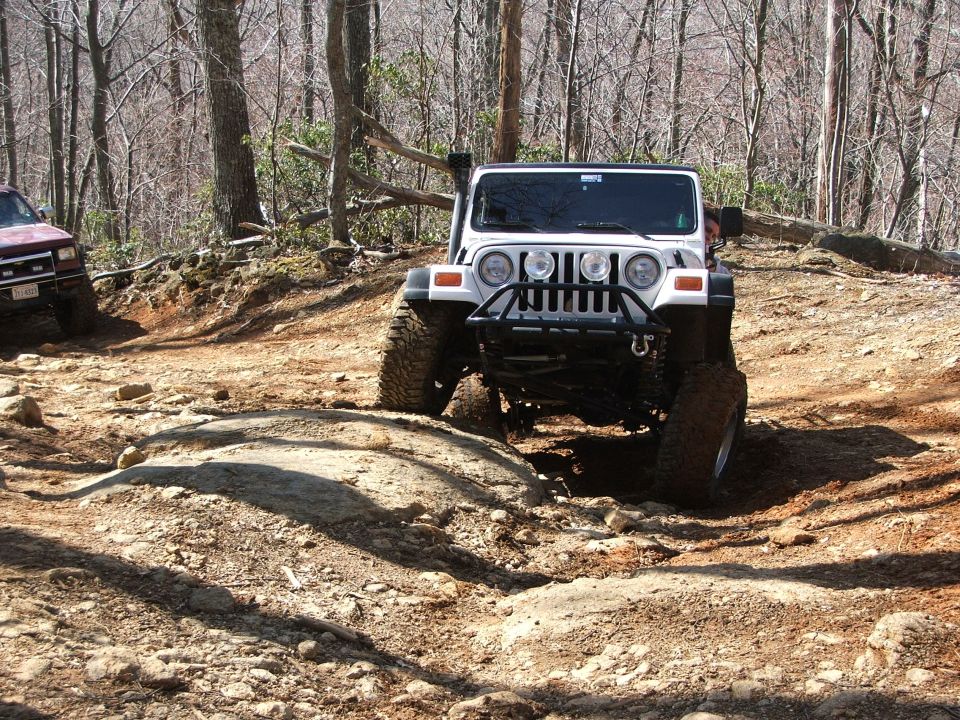 White Jeep Wrangler climbing a rocky trail, maroon SUV in background