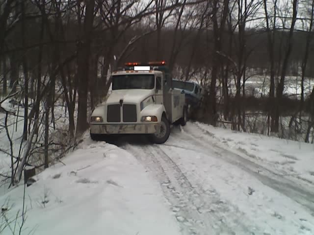 White tow truck with orange lights on top, towing a blue car on a snowy road
