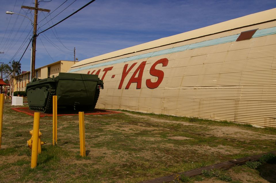 Green amphibious tracked vehicle parked on display in front of a corrugated metal building with red lettering