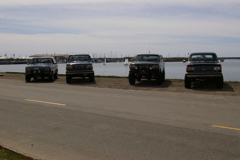 1986 Ford Bronco - Several Ford trucks are parked along the waterfront, with a harbor and sailboats visible behind them.
