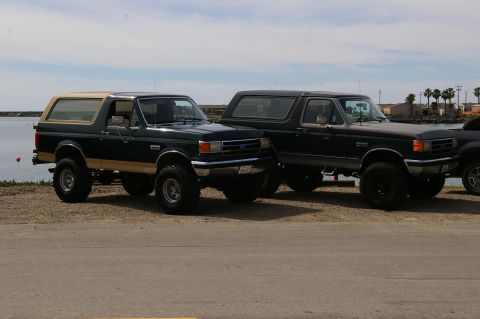 1986 Ford Bronco - Two Ford Broncos are parked next to each other near a body of water under a partly cloudy sky.