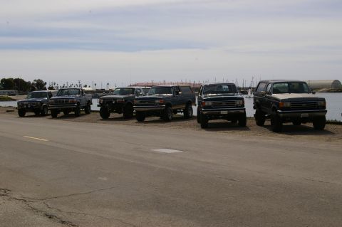 1986 Ford Bronco - A lineup of Ford Bronco SUVs and pickup trucks are parked near a body of water under a cloudy sky.