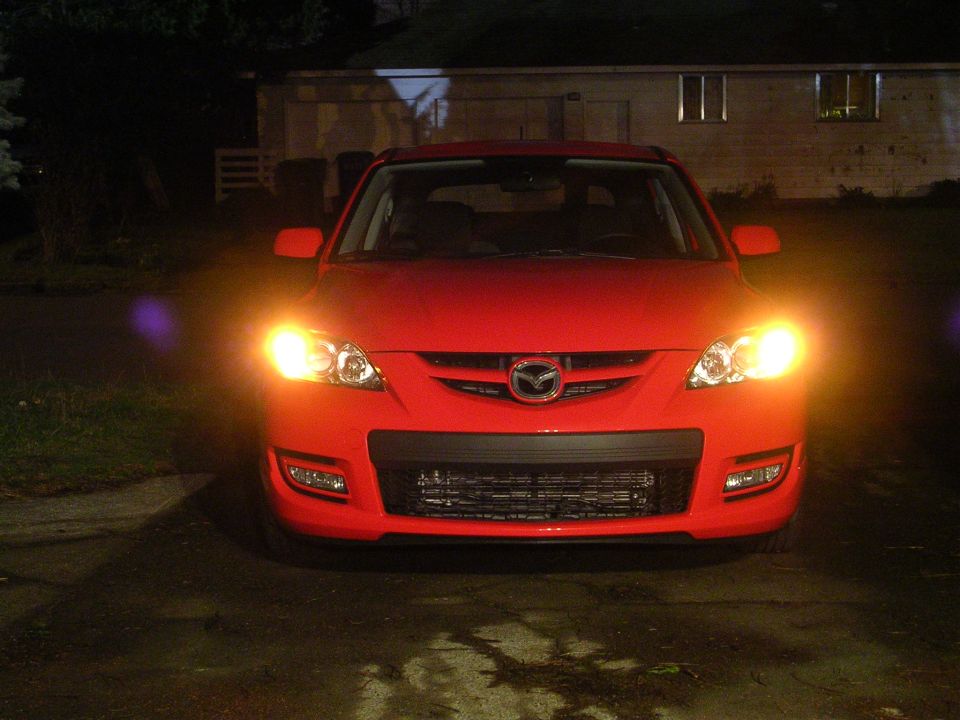 Red Mazda 3 hatchback front view at night with lights on