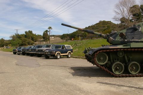 1986 Ford Bronco - A military tank is parked in a lot next to several trucks and SUVs under a cloudy sky.