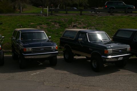 1986 Ford Bronco - Two Ford Bronco SUVs are parked in a lot, with a third vehicle partially visible on the right.