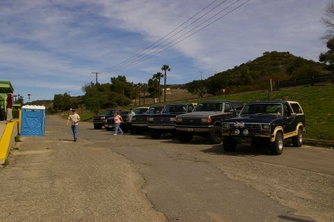 1986 Ford Bronco - A group of Ford trucks and Broncos are parked on a paved area with a hill in the background.