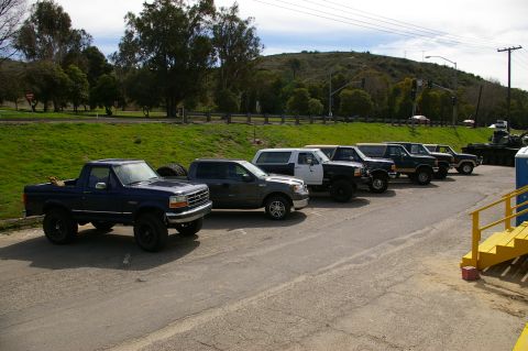1986 Ford Bronco - A line of trucks and SUVs are parked in a lot, with a dark blue SUV in the foreground.
