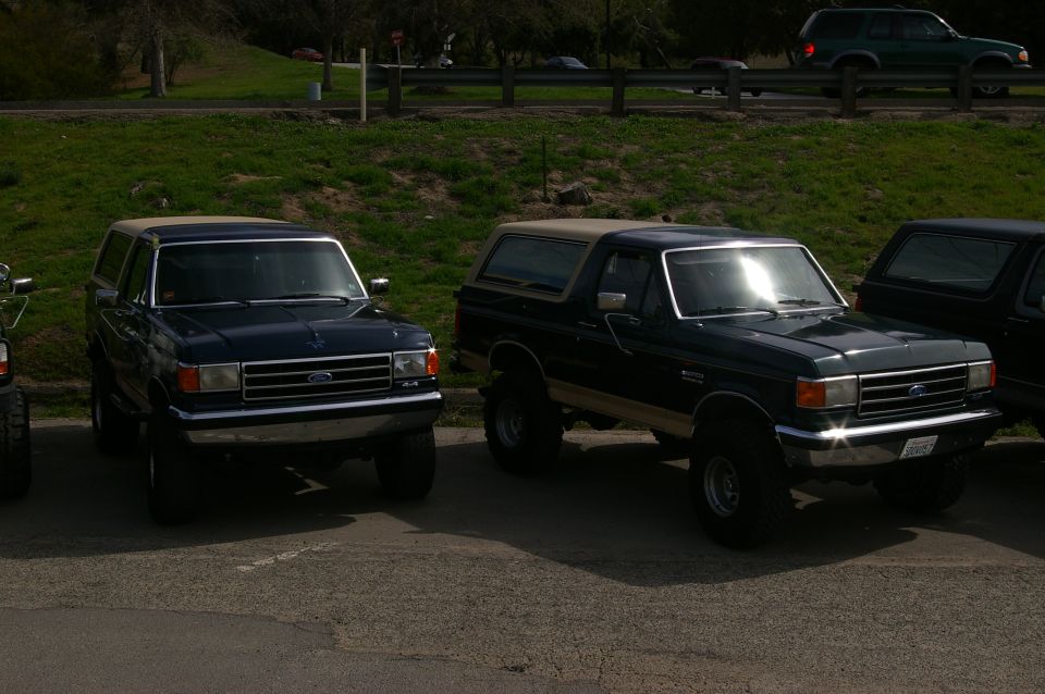 Two Ford Broncos parked side-by-side. The one on the left is dark blue, the one on the right is dark green with a tan stripe