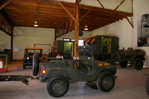 1986 Ford Bronco - A green military jeep with USMC 308404 markings is displayed indoors alongside other vehicles and equipment.