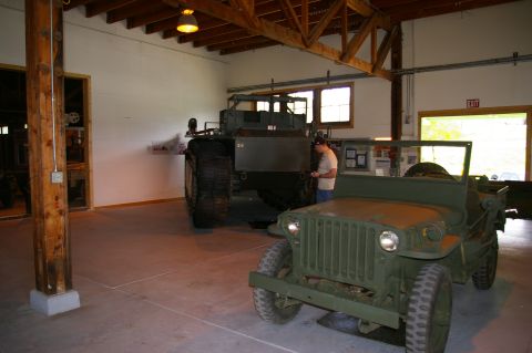 1986 Ford Bronco - Two military vehicles are parked inside a large garage, one in the foreground and another larger one behind it.