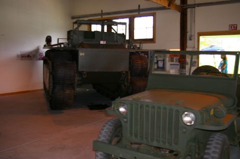 1986 Ford Bronco - Two military vehicles are displayed indoors, one in the foreground and a larger one behind it.