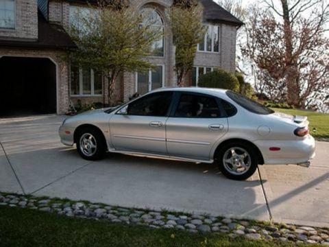 1997 Ford Taurus - A silver sedan with a spoiler is parked on a driveway in front of a large brick house.