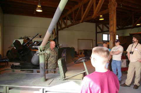 1986 Ford Bronco - A group of people observe a military vehicle with a mounted weapon inside a large structure.